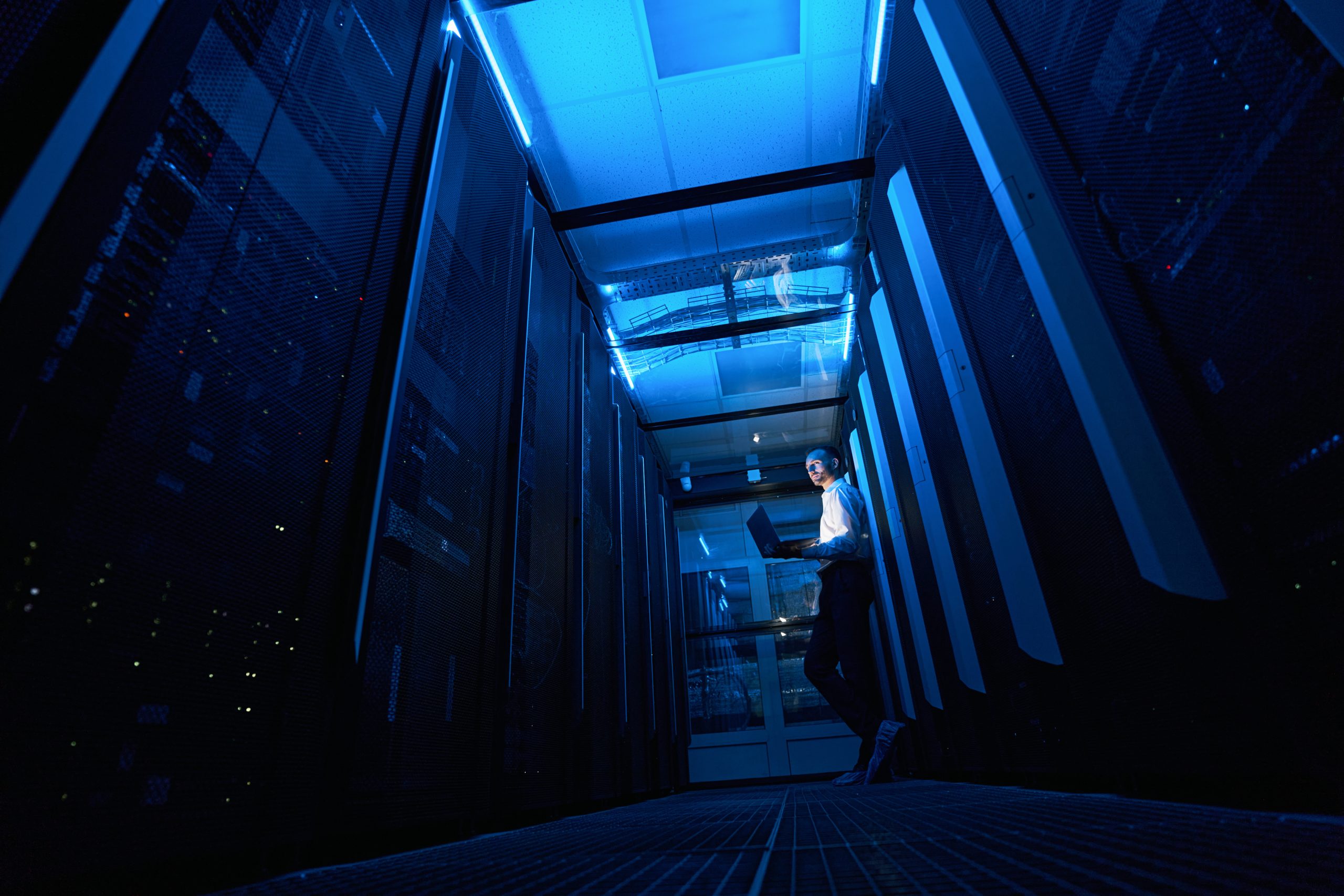 Data center technician resting against network equipment in server room aisle with laptop